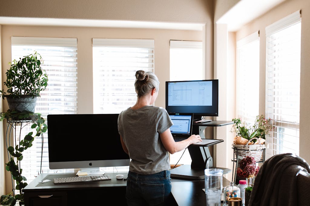 A woman standing while using a laptop and monitor in a bright, modern home office space.