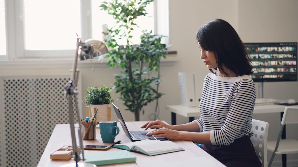 Focused young woman working on a laptop in a bright office setting with plants and desk accessories.
