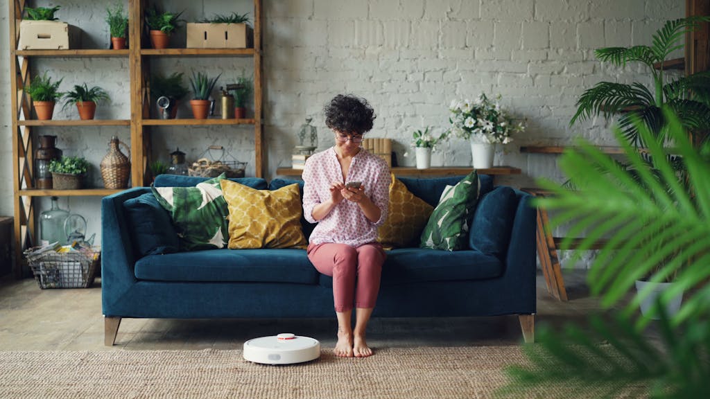 Woman sits on sofa using smartphone while robotic vacuum cleans living room floor.
