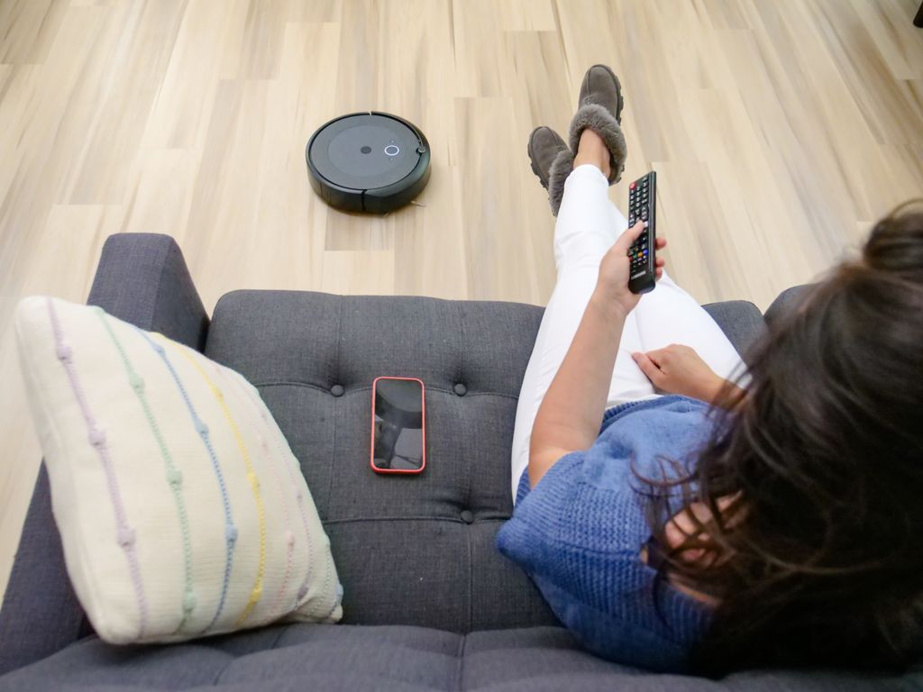 Person relaxing on a sofa controlling a robot vacuum cleaner with a remote.
